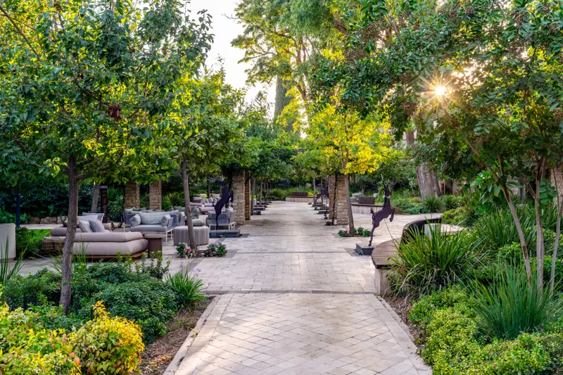 Wide stone garden path between green trees and plants in soft evening sun at Ayala by Isrotel Collection