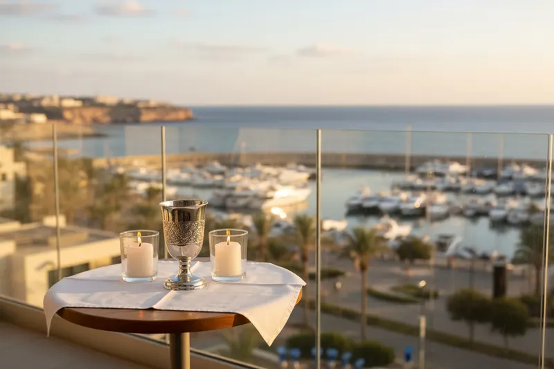 Hotel balcony at golden hour above a marina and the sea, a silver kiddush cup and two candles in glass holders on a table